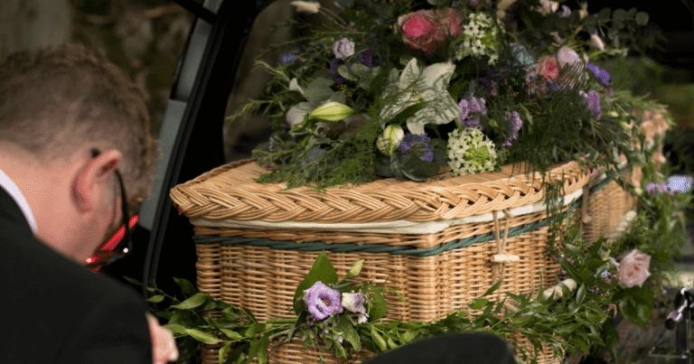 Close-up view of a wicker casket heavily decorated with mixed white, pink, and purple flowers and foliage, as a person in a black suit looks on