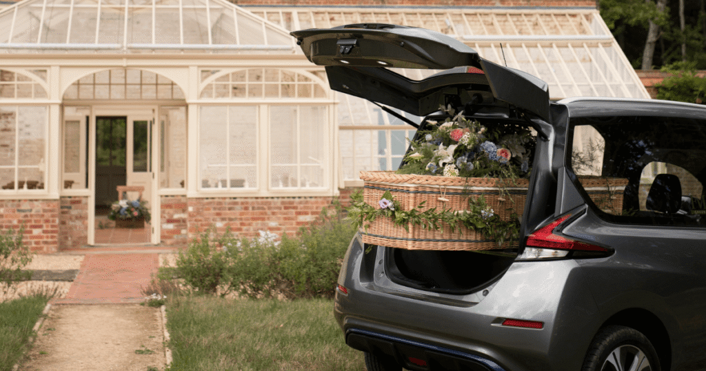 The open trunk of a hearse reveals a wicker casket laden with flowers, parked on a grassy area in front of a brick building with a glass conservatory entrance
