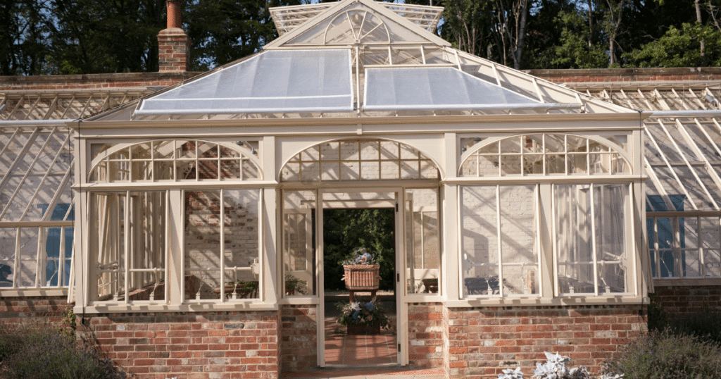 An elegant Victorian-style conservatory or greenhouse with a red brick base, large glass panes, and an open central door where a flower basket is visible