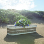 The Seagrass Coffin on the sand with large purple, pink and white flower spray on top