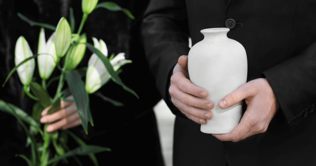 Close-up of a person in a black suit holding a plain white ceramic cremation urn, with another person nearby holding white lily flowers